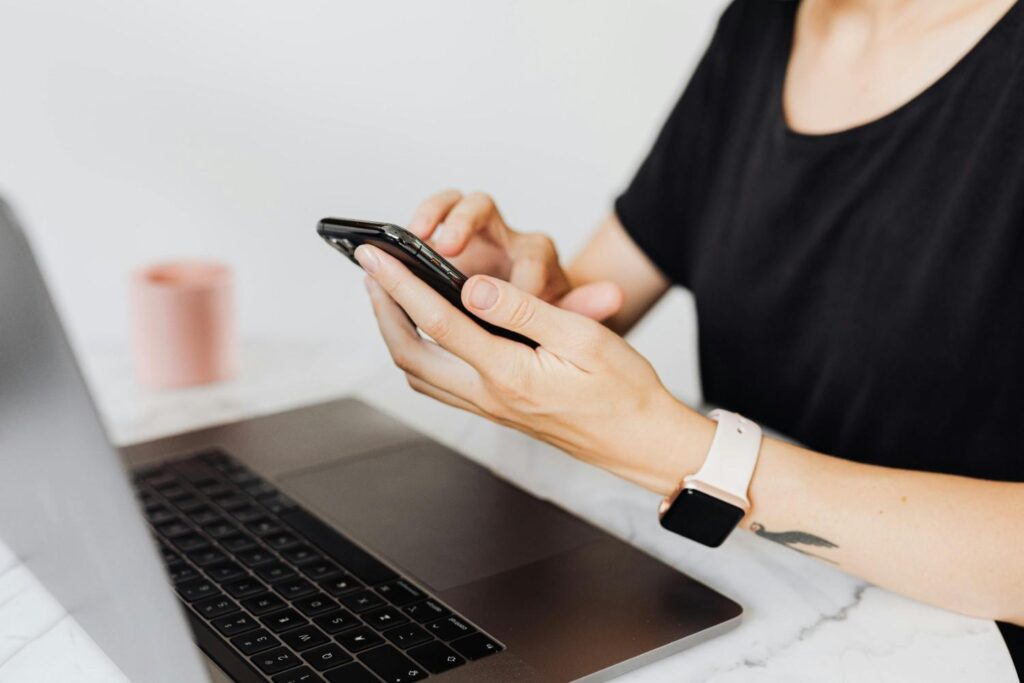 A woman using a smartphone with a laptop on an office desk